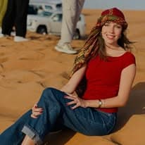 Guest in a traditional red headscarf resting on a red dune, safari 4x4s parked on the ridge behind